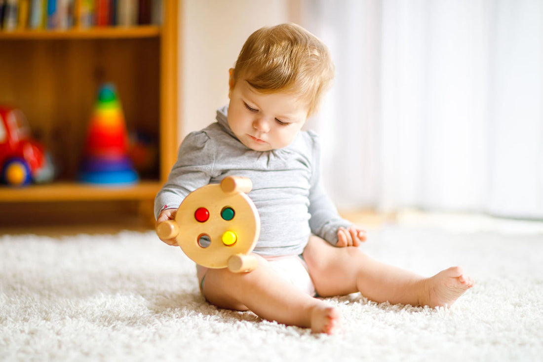 A baby in a gray outfit sits on a soft carpet, holding a colorful wooden toy. In the background, a blurry bookshelf and toys add warmth to the sunny room.