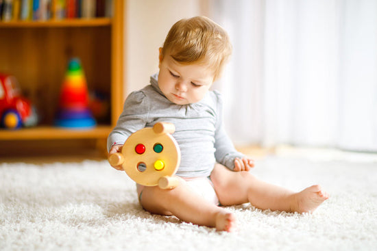A baby in a gray outfit sits on a soft carpet, holding a colorful wooden toy. In the background, a blurry bookshelf and toys add warmth to the sunny room.