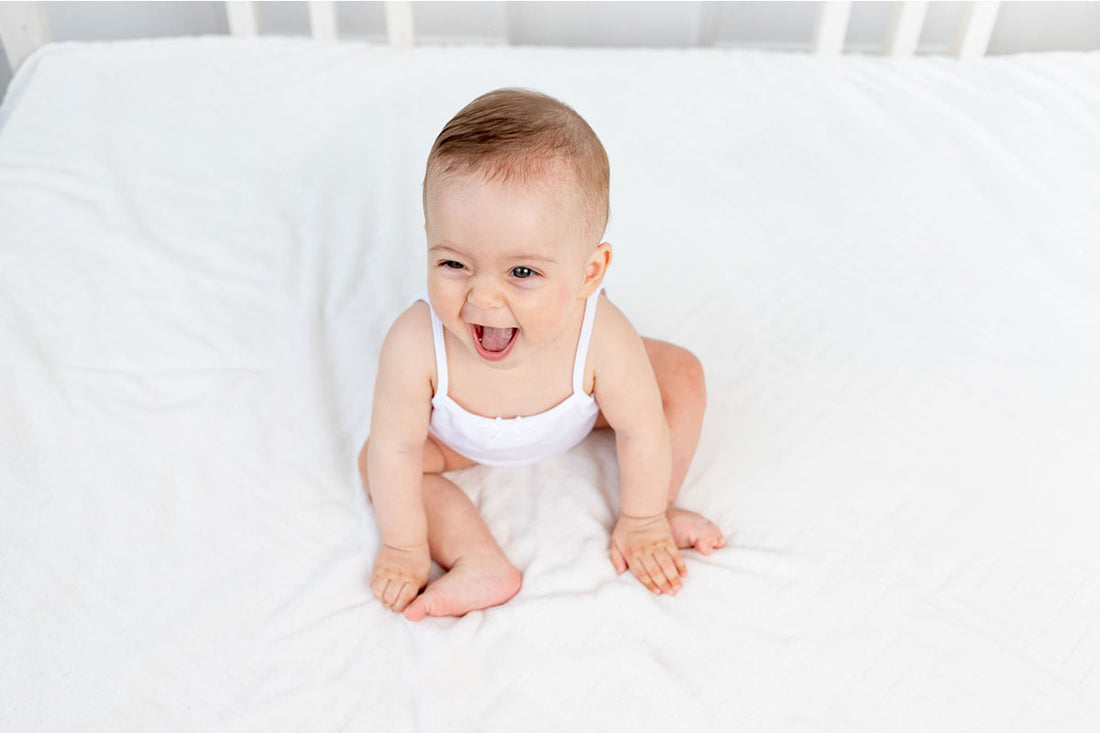 A joyful baby in a white onesie sits on a white blanket, smiling and looking upward with excitement. The scene conveys happiness and warmth.