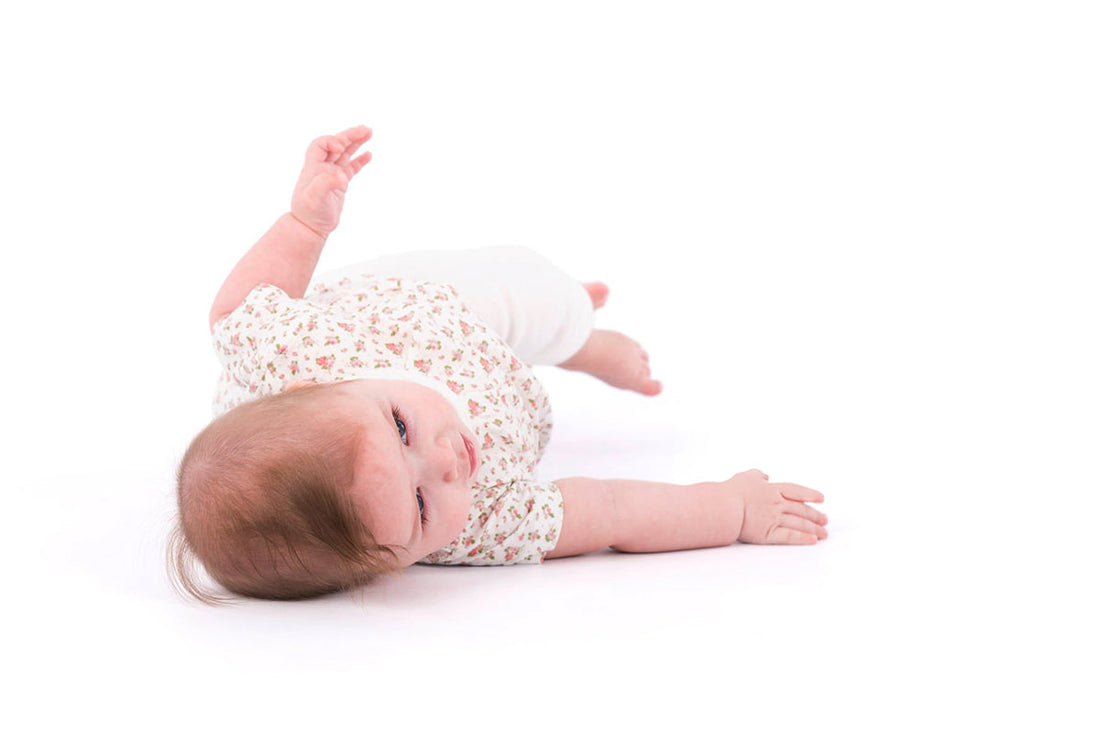 A baby lying on their side on a white background, wearing a small floral-patterned shirt and white pants, with one arm raised and looking slightly toward the camera as if mid-roll.