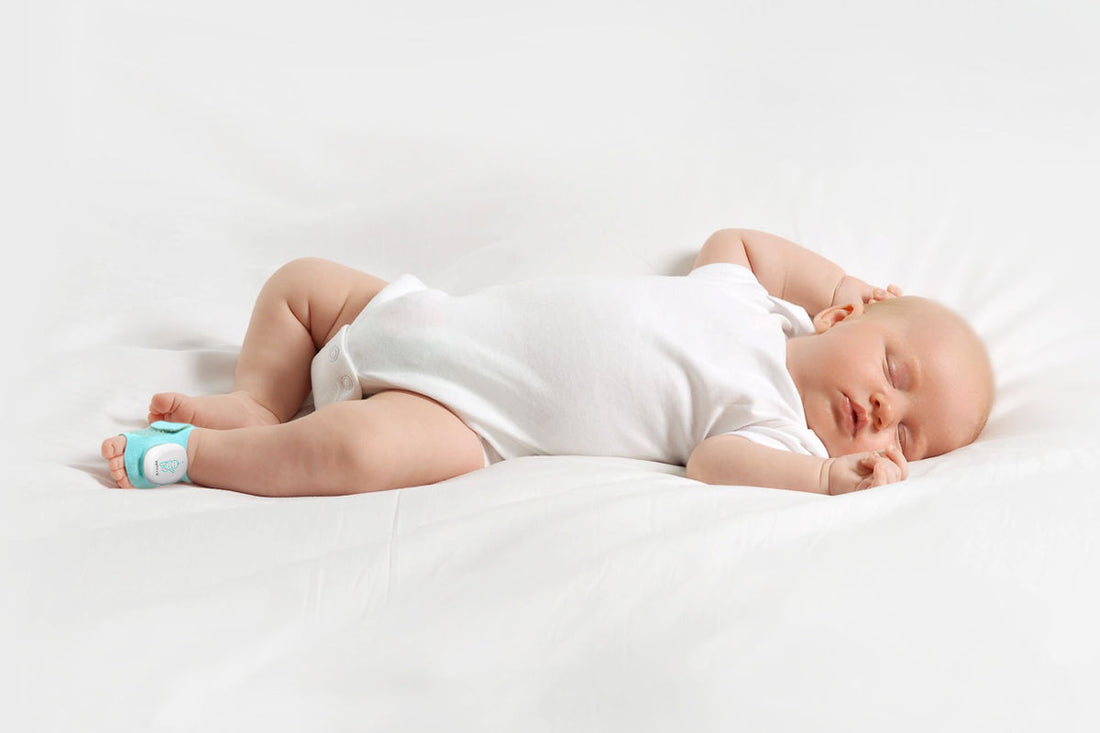 Sleeping baby lying on their side on a white bed, wearing a Sense-U smart sock shoe on one foot.