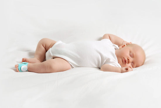 Sleeping baby lying on their side on a white bed, wearing a Sense-U smart sock shoe on one foot.