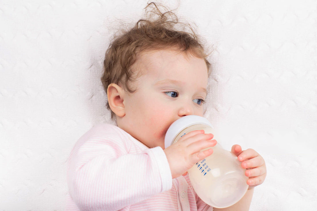 A baby in a pink striped onesie drinking from a baby bottle while lying on a soft white surface, holding the bottle with both hands.