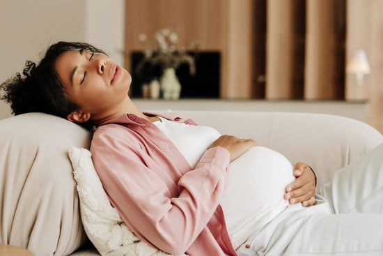 Pregnant woman resting on a sofa with eyes closed, gently holding her belly, expressing pregnancy fatigue and the need for rest in a calm home environment.