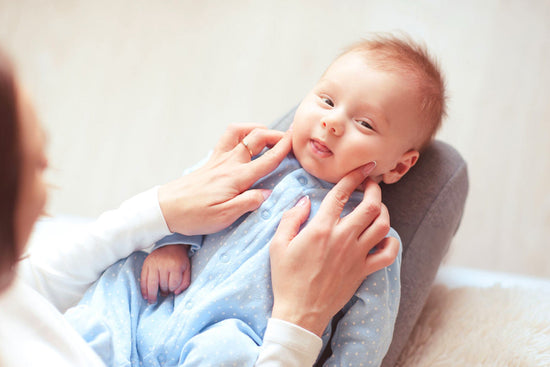 A baby lying comfortably while an adult gently touches their cheeks, the baby smiling softly in a calm, bright setting.