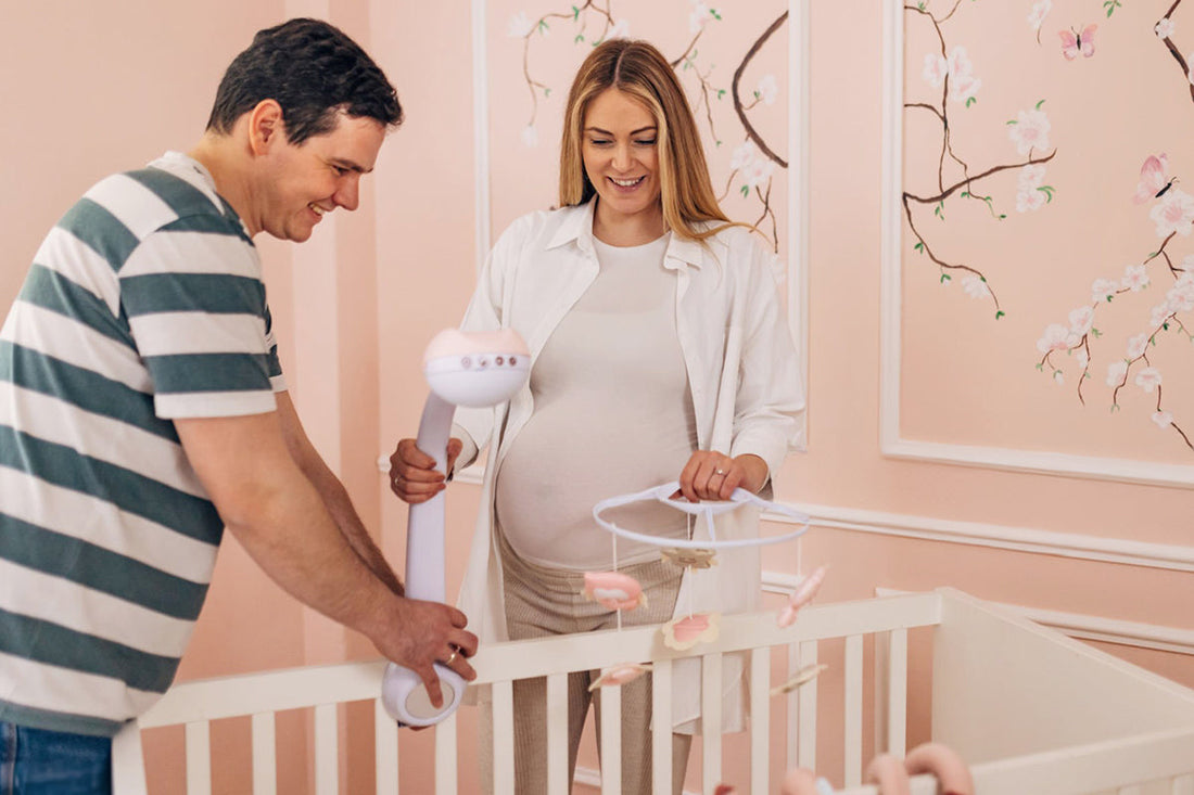 A pregnant woman and a man smile while setting up a mobile on a white crib in a pink, floral-decorated nursery.