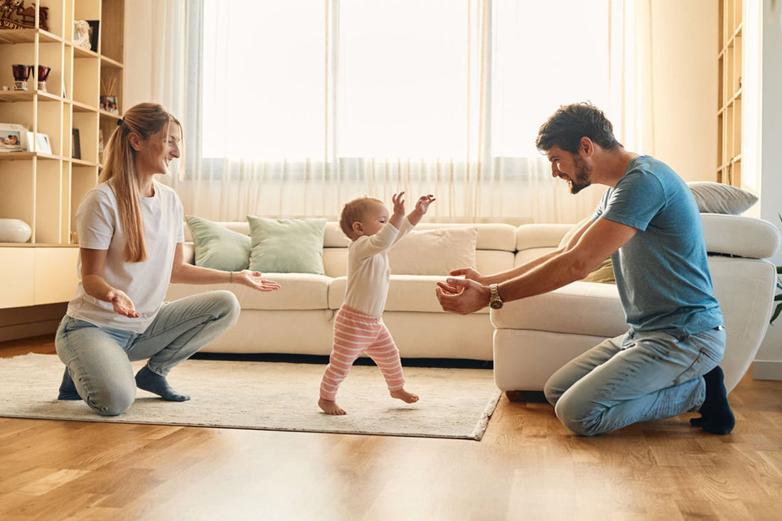A baby takes first steps towards a smiling father, while the mother encourages nearby. The scene is warm and joyful in a bright, cozy living room.