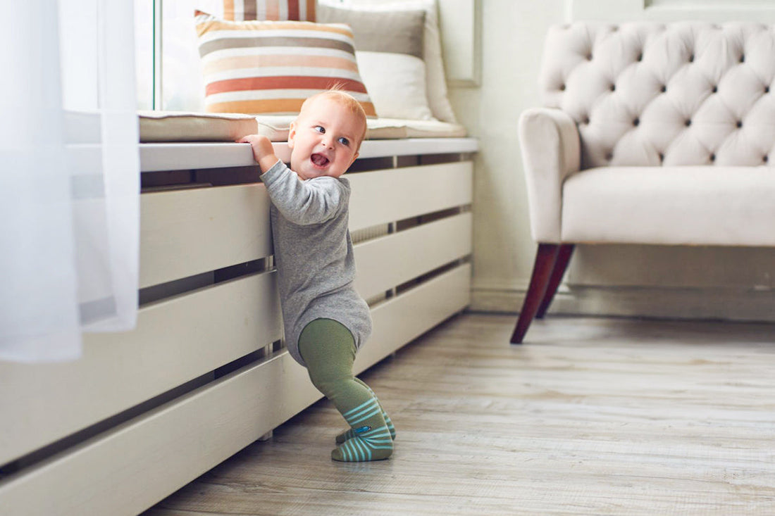 A cheerful baby in a gray outfit stands holding onto a window seat, wearing striped socks. The room features a cushioned bench and a beige armchair.