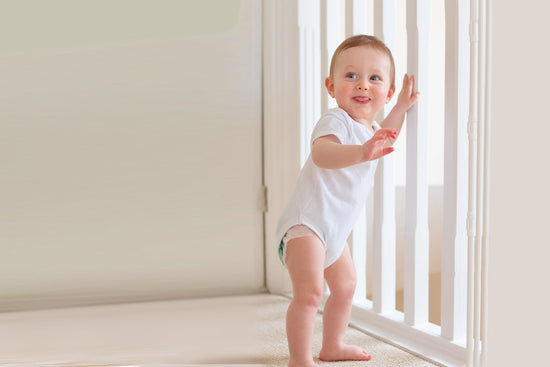 A smiling baby in a white onesie stands holding onto a white safety gate indoors. The setting feels safe and joyful, suggesting a milestone moment.