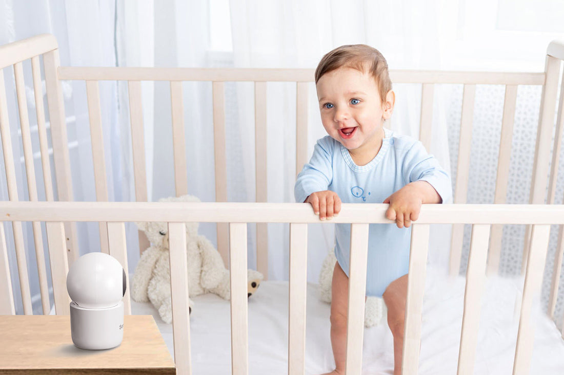 A happy baby in a light blue onesie stands in a crib, smiling. A plush teddy bear is in the crib, and a white baby monitor sits on a nearby table.