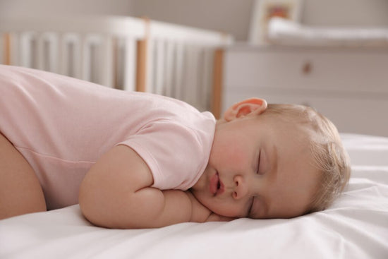 A baby in a pink onesie sleeping on their side in a crib, resting on a white sheet with hands tucked under their cheek