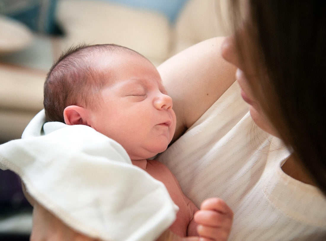Sleeping newborn baby rests in a mother’s arms, wrapped in a white blanket; their close embrace conveys warmth, tenderness, and calm.