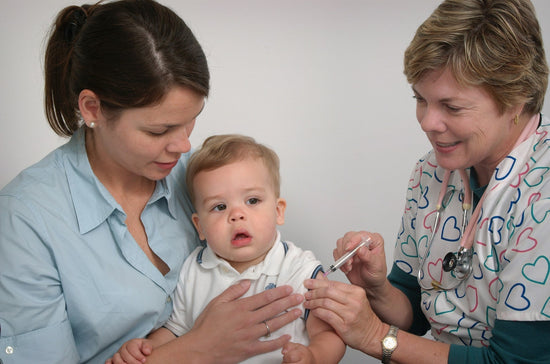 A healthcare worker gives a vaccination to a young child held by an adult woman. The scene takes place in a medical setting with a neutral background.