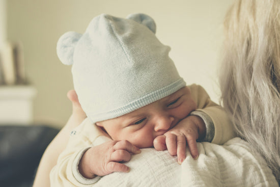 A newborn baby, wearing a blue hat with bear ears, rests on an adult's shoulder, exuding warmth and comfort in a softly lit environment.
