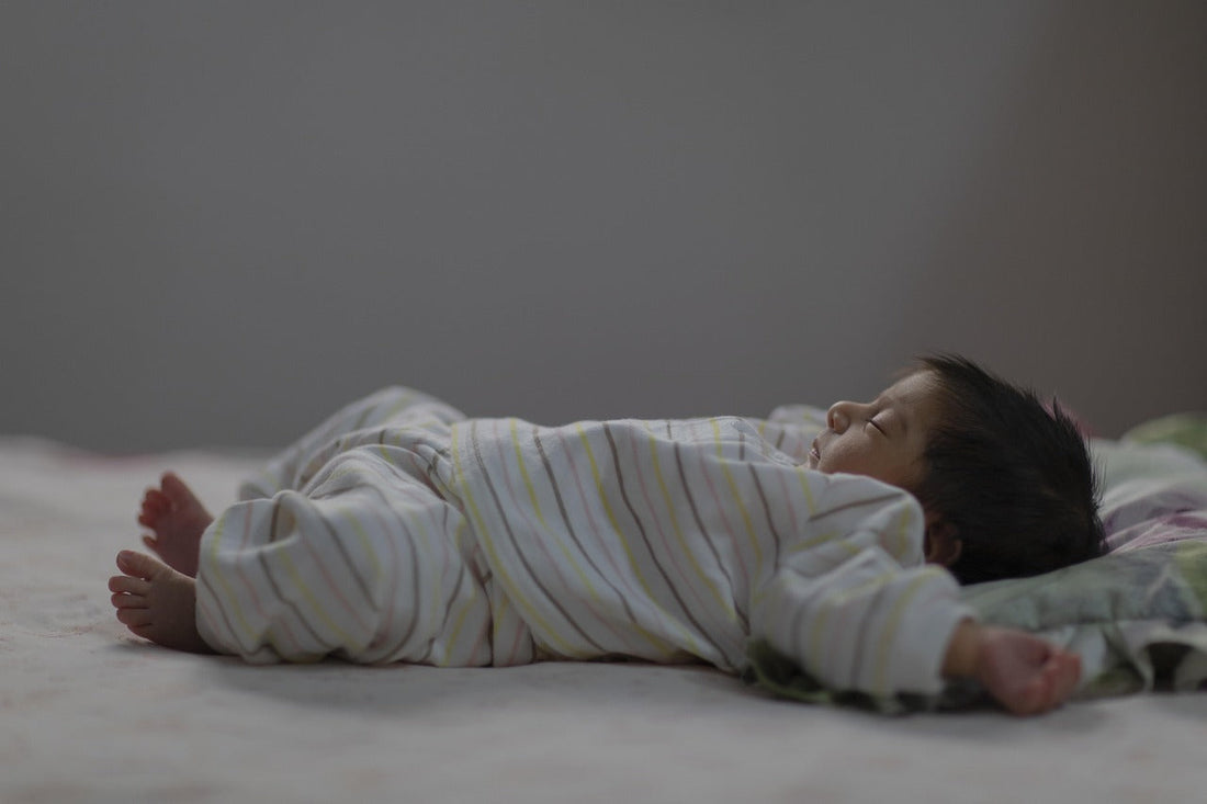 Sleepy baby in striped pajamas lies face down across a bed, arms outstretched and feet visible, creating a quiet, tender mood.