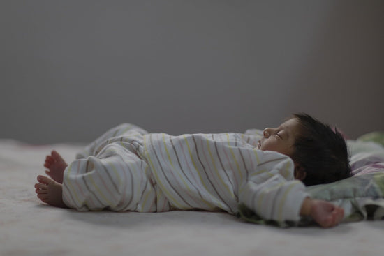 Sleepy baby in striped pajamas lies face down across a bed, arms outstretched and feet visible, creating a quiet, tender mood.