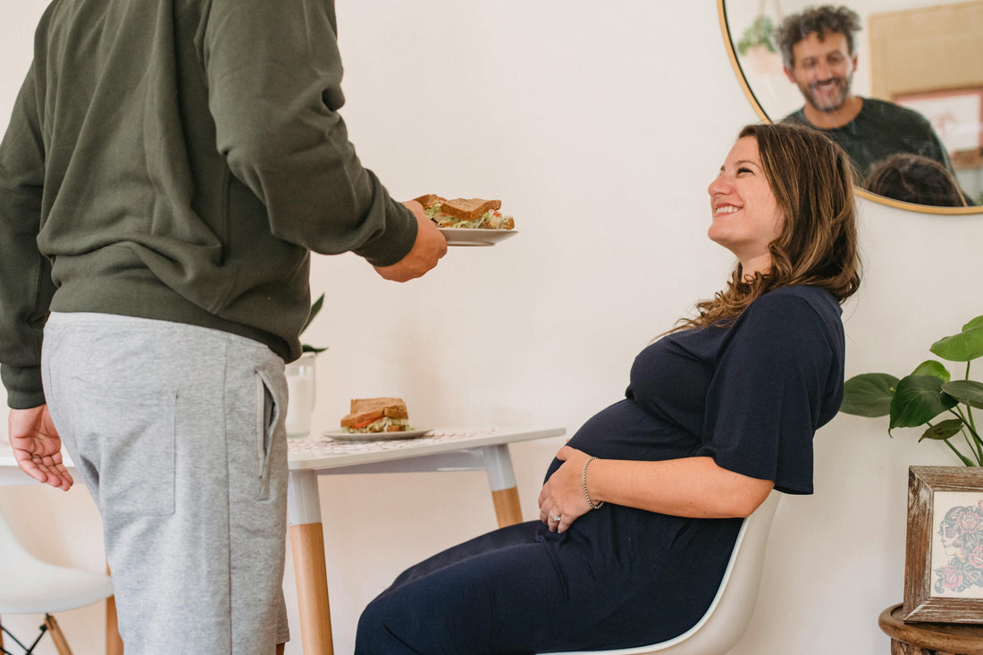 Smiling pregnant woman seated at a table as a man serves her a sandwich. A mirror reflects both, creating a warm and caring atmosphere.
