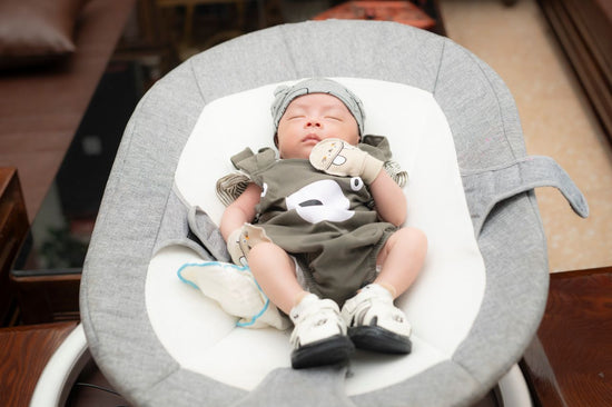 Sleeping newborn baby in a gray bouncer, wearing a hat, mittens, and outfit, with legs raised and a peaceful expression.