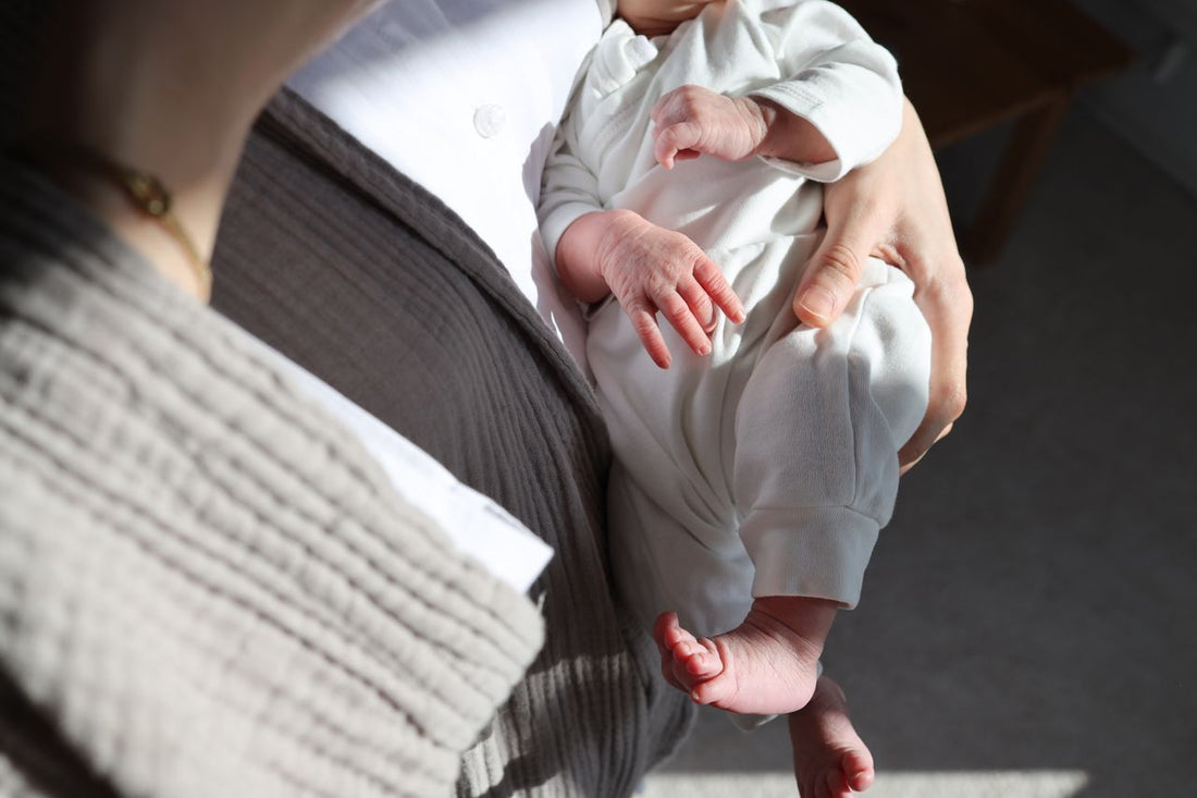 Close-up of an adult gently holding a newborn in a white onesie, with tiny hands and feet visible in warm sunlight; tender, joyful mood.