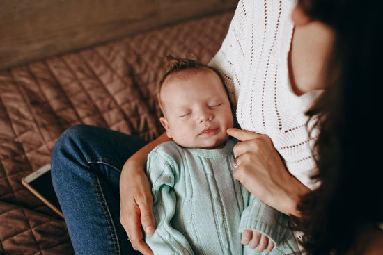 A sleeping baby in a pale green sweater rests on a woman’s lap as she gently touches the baby’s cheek on a cozy brown sofa, creating a tender mood.