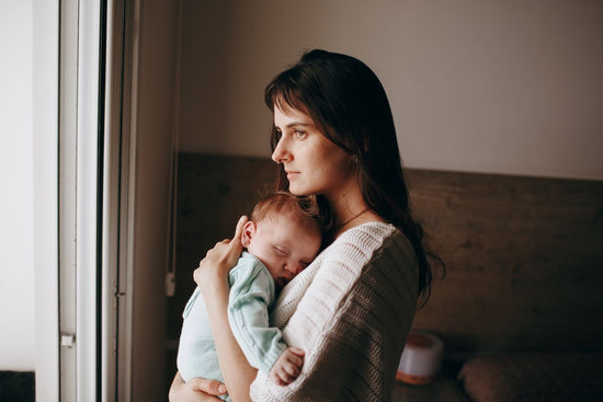 Woman holding a sleeping baby looks out a window, standing in a softly lit room with neutral colors.