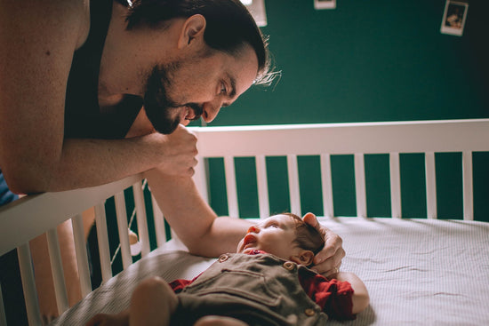 A bearded man leans over a white crib, gazing lovingly at a baby lying inside. The baby, in a red outfit, looks back peacefully. Soft, warm lighting creates a tender atmosphere.