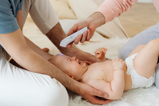 A baby lying on a soft surface is having its temperature taken with a forehead thermometer. Two adults gently support the baby, showing care and attention.
