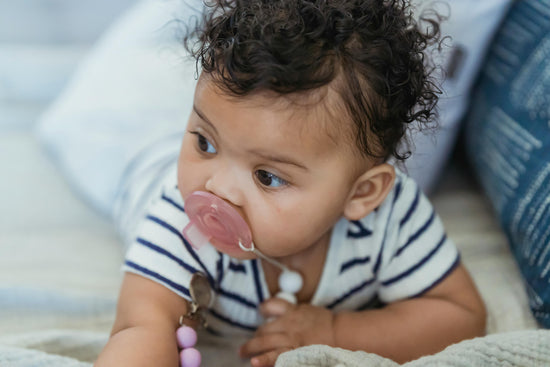 A baby with curly hair and a striped onesie looks curiously to the side, pacifier in mouth, lying on a soft, beige blanket. Calm and inquisitive tone.