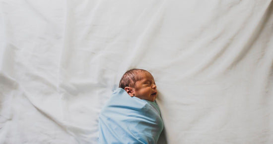 A newborn with eyes closed, wrapped snugly in a soft blue blanket on rumpled white bedding, creates a quiet, tender, peaceful scene.