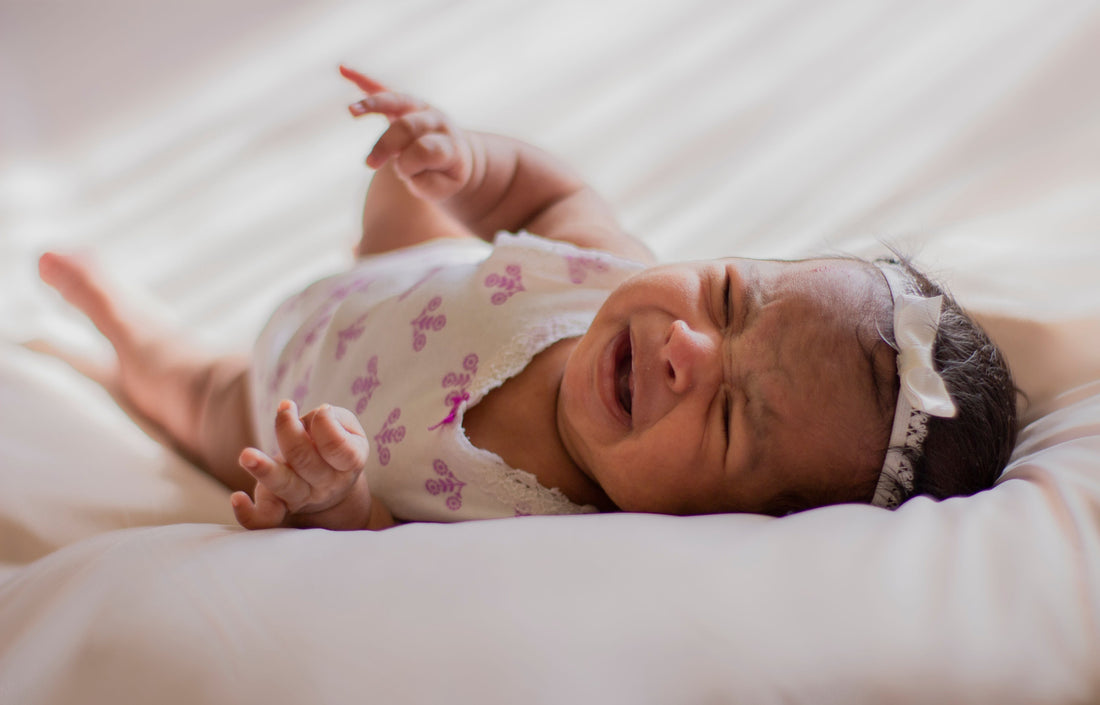 Newborn baby girl in a floral onesie and white headband lies on a soft white bed, eyes closed and crying, arms raised in distress.