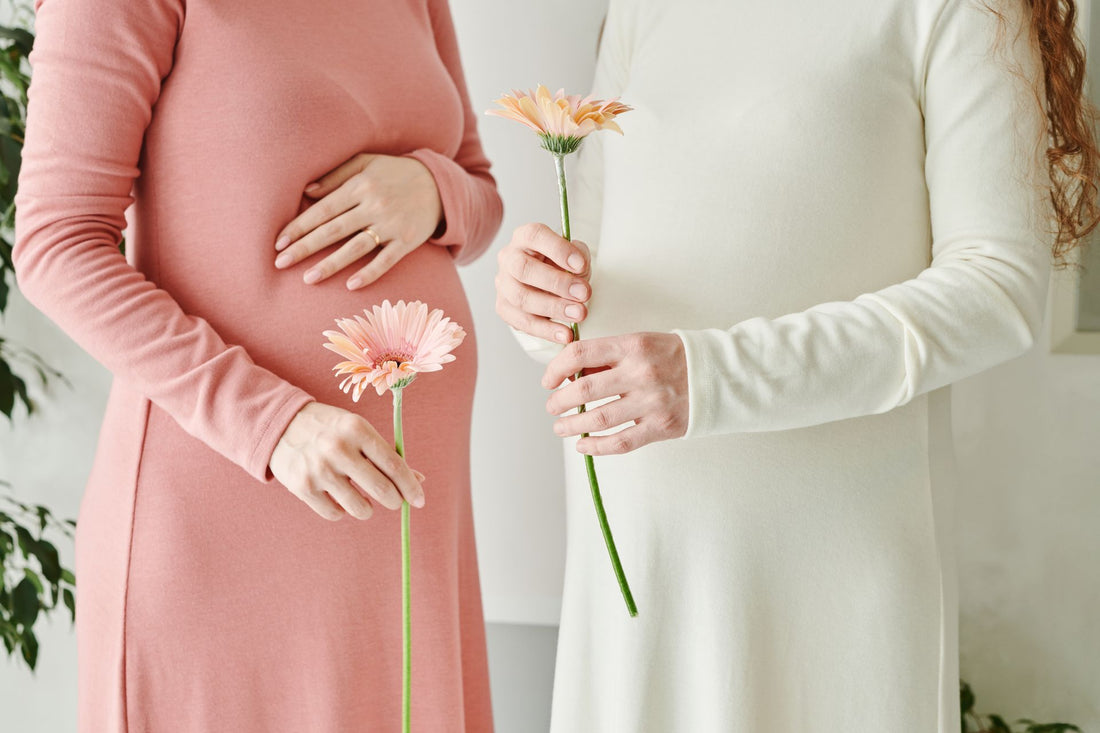 Two pregnant women standing side by side, one in a pink dress and one in white, each holding a single pink gerbera daisy against their baby bumps.