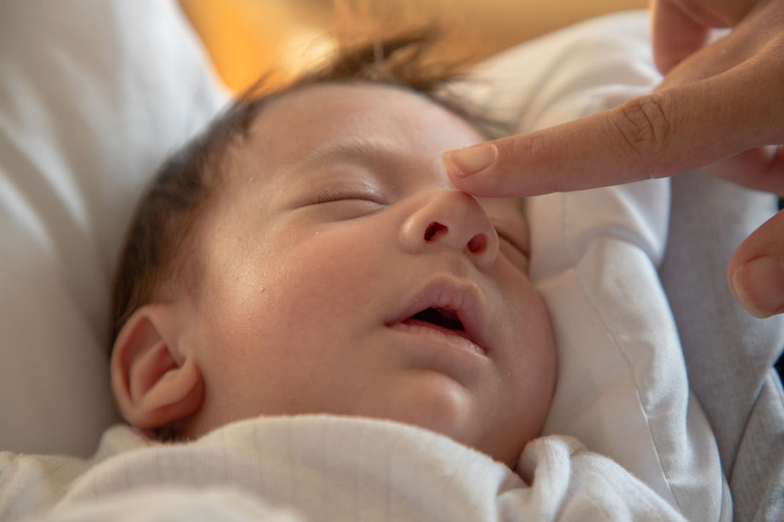Sleeping newborn baby wrapped in a white blanket, eyes closed, as an adult gently touches the baby’s nose in a tender moment.