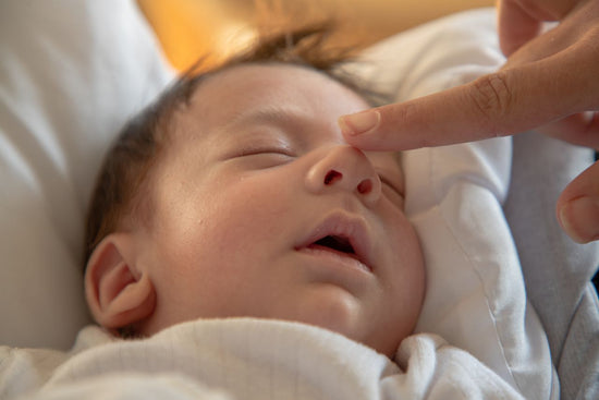 Sleeping newborn baby wrapped in a white blanket, eyes closed, as an adult gently touches the baby’s nose in a tender moment.