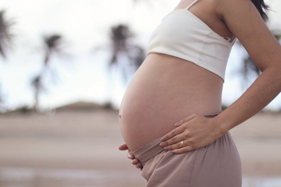 Pregnant woman in a white top and tan skirt caressing her belly on a beach with blurred palm trees in the background, conveying serenity.