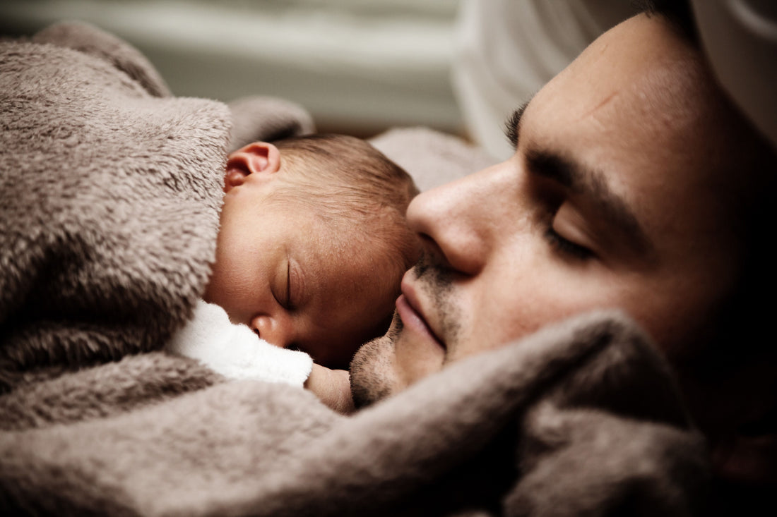 A newborn baby sleeps peacefully on a man's chest, both wrapped in a soft brown blanket. The scene conveys warmth, tenderness, and comfort.