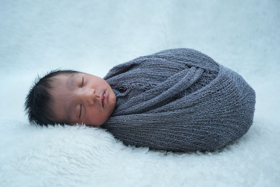 A newborn baby sleeps peacefully, wrapped snugly in a soft gray blanket. The background is a fluffy white texture, conveying warmth and comfort.