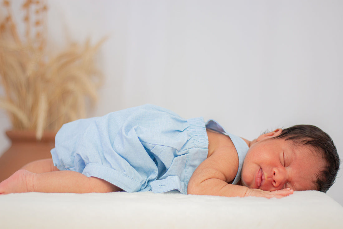 Newborn baby wearing a light blue outfit is lying on their stomach, sleeping peacefully on a soft white surface.
