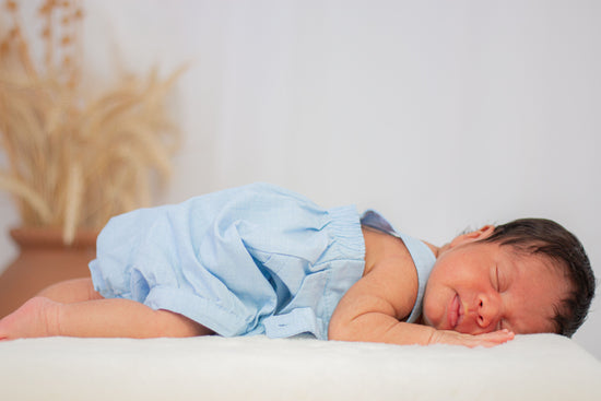 Newborn baby wearing a light blue outfit is lying on their stomach, sleeping peacefully on a soft white surface.
