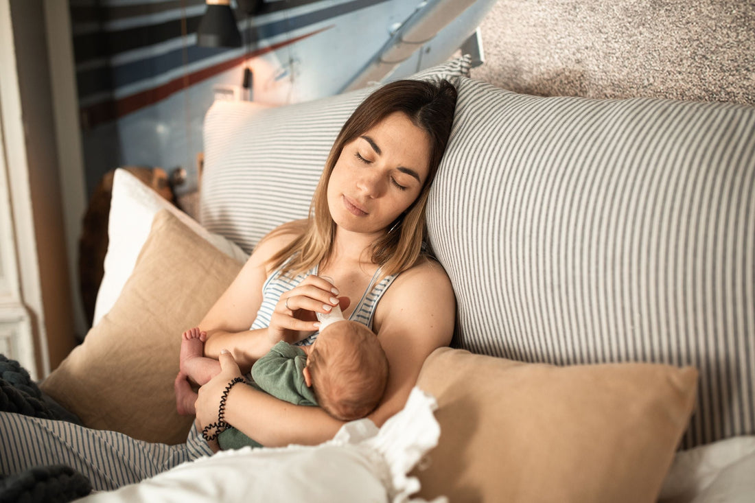 A woman peacefully bottle-feeds a baby while lounging on a bed. The room is cozy, with soft lighting and striped pillows, creating a serene atmosphere.