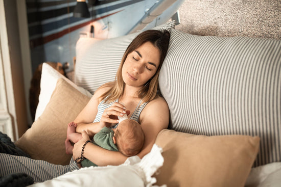 A woman peacefully bottle-feeds a baby while lounging on a bed. The room is cozy, with soft lighting and striped pillows, creating a serene atmosphere.