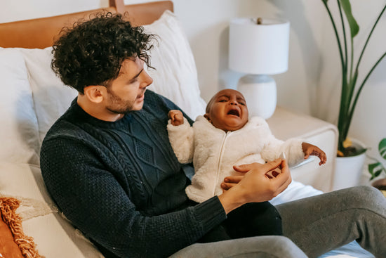 Father cradles a crying baby in a cozy bedroom, offering comfort on a bed beside a lamp and houseplant; tender family moment.