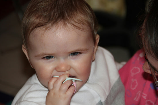 A baby with short, light brown hair is nibbling on a spoon while being held. The baby has a focused expression, and a caregiver in colorful clothing is nearby.